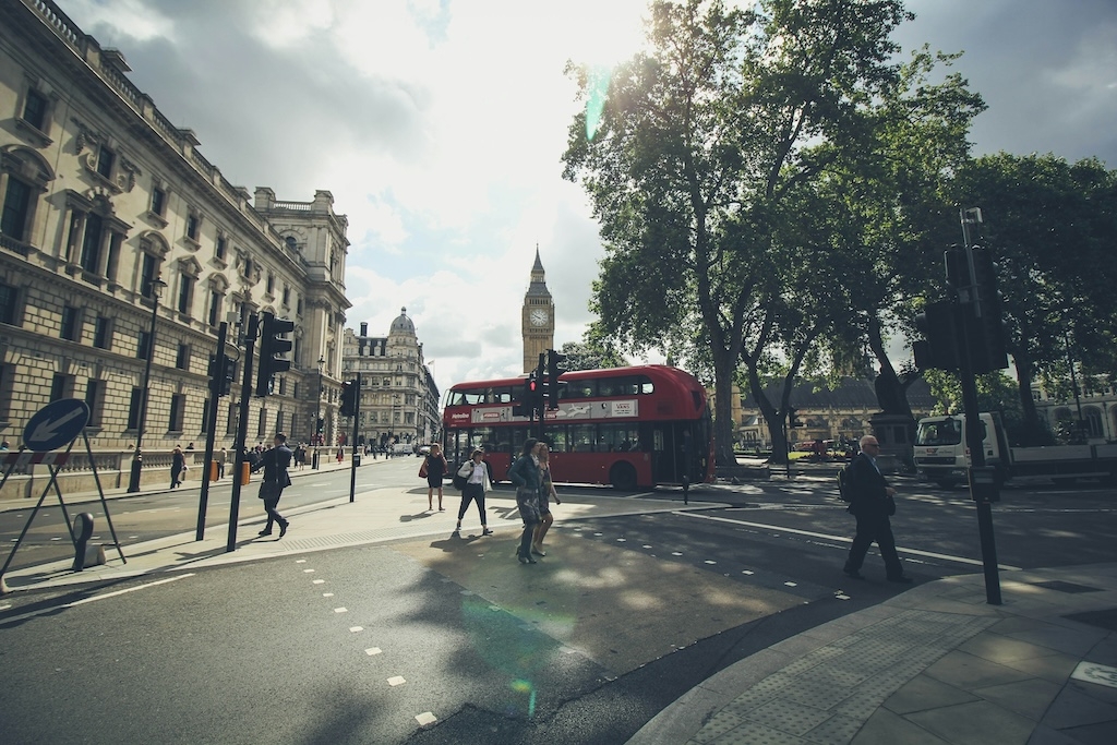 Photo of a London street taken during a sunny day. Pedestrians walk across a street with a red double-decker bus passing by. In the distance, Big Ben can be seen.